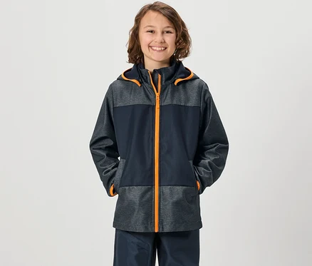 Lächelnder Junge in Kinder-Softshelljacke mit Händen in den Taschen.
