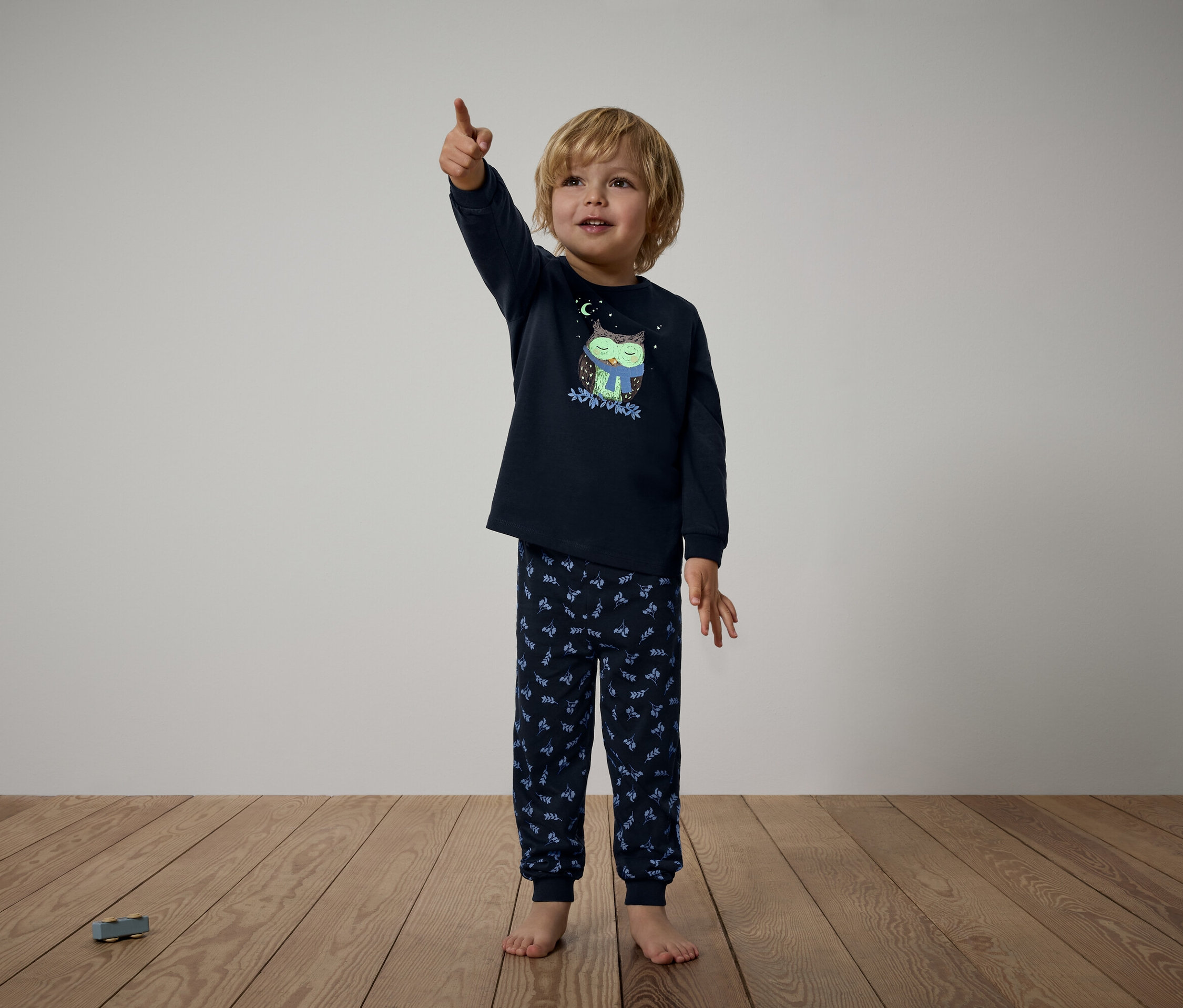 Junge im dunkelblauen Pyjama mit Eulenmuster zeigt mit dem Finger nach oben.