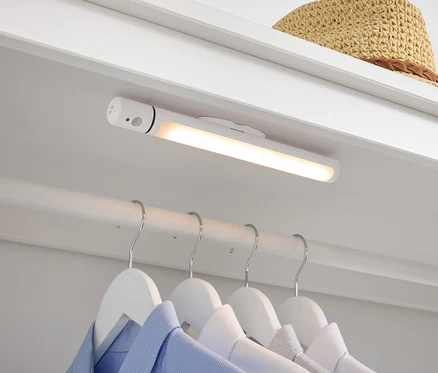 Im Kleiderschrank befinden sich eine weiße LED-Lampe, darunter Hemden auf weißen Kleiderbügeln und oben liegt ein Strohhut.