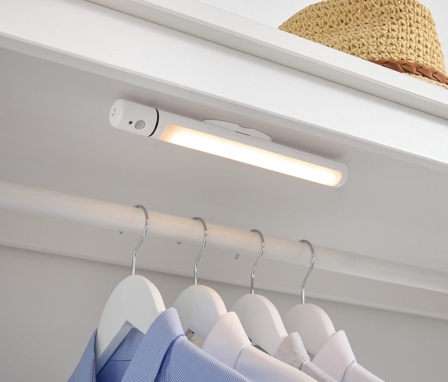 Im Kleiderschrank befinden sich eine weiße LED-Lampe, darunter Hemden auf weißen Kleiderbügeln und oben liegt ein Strohhut.