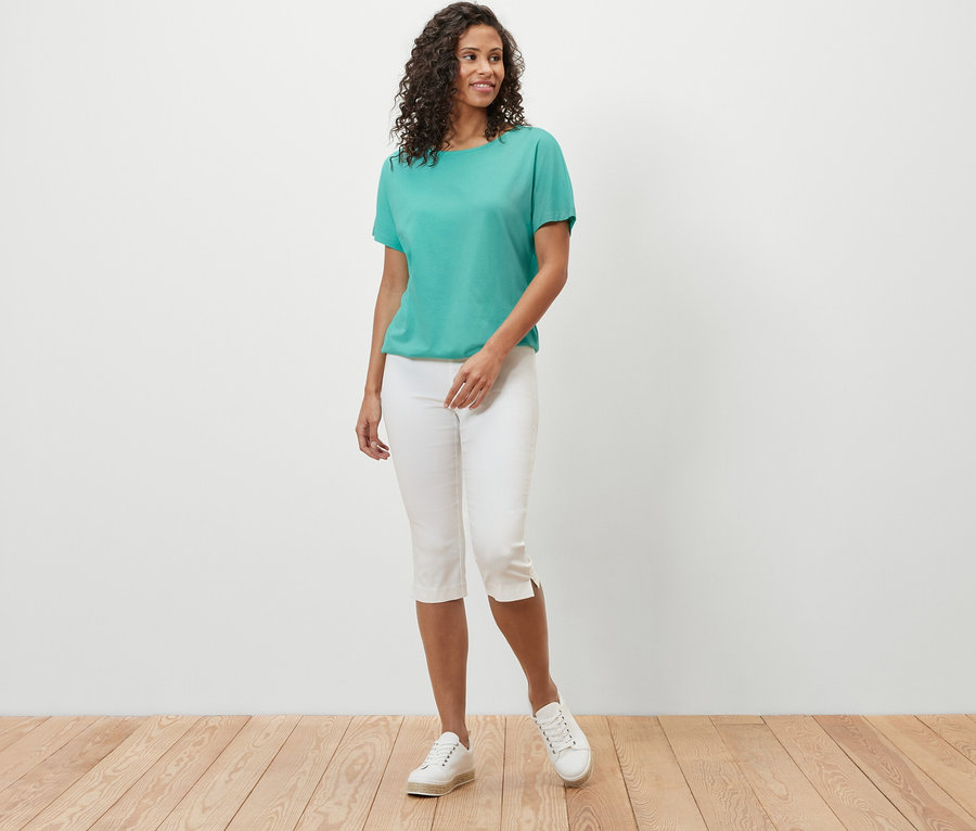 Eine Frau posiert in einem aqua überschnittenen Blusenshirt und weißen Caprihosen.