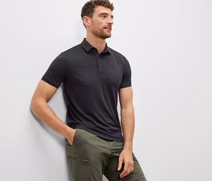 Mann lehnt an einer Wand und trägt ein anthrazitfarbenes Funktions-Poloshirt.