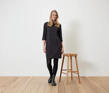 Frau steht neben einem Holzhocker und trägt ein Hahnentrittkleid, schwarze Leggings und Stiefel.
