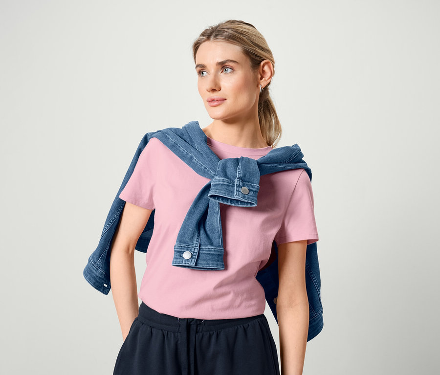 Frau mit Jeansjacke über den Schultern, trägt ein rosafarbenes Basic T-Shirt und Bermuda-Sweatshorts.