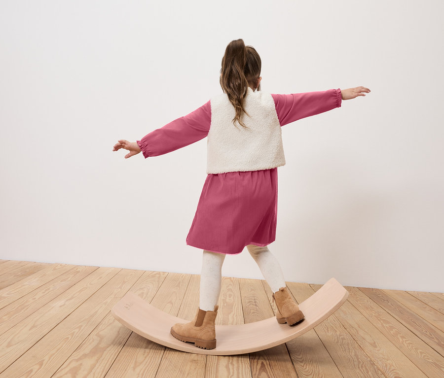 Mädchen in rosa Kleid, Weste und braunen Stiefeln balanciert auf einem hölzernen Balance Board.