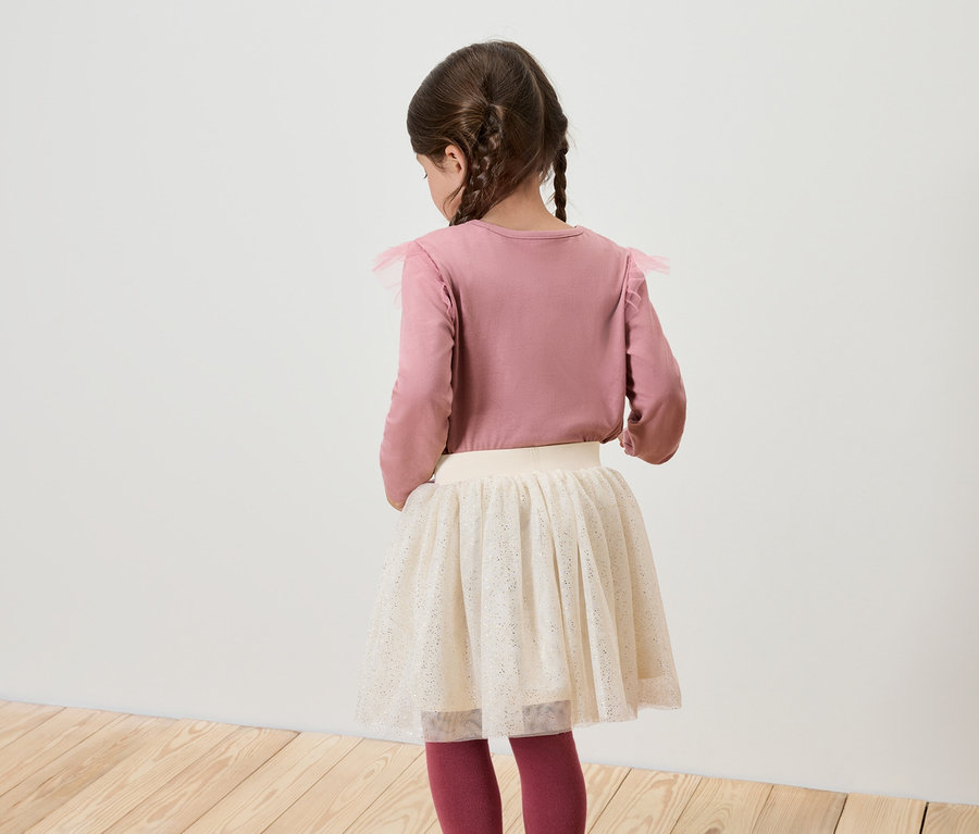 Mädchen mit dem Rücken zur Kamera trägt ein rosa Langarmshirt und einen weißen Rock.