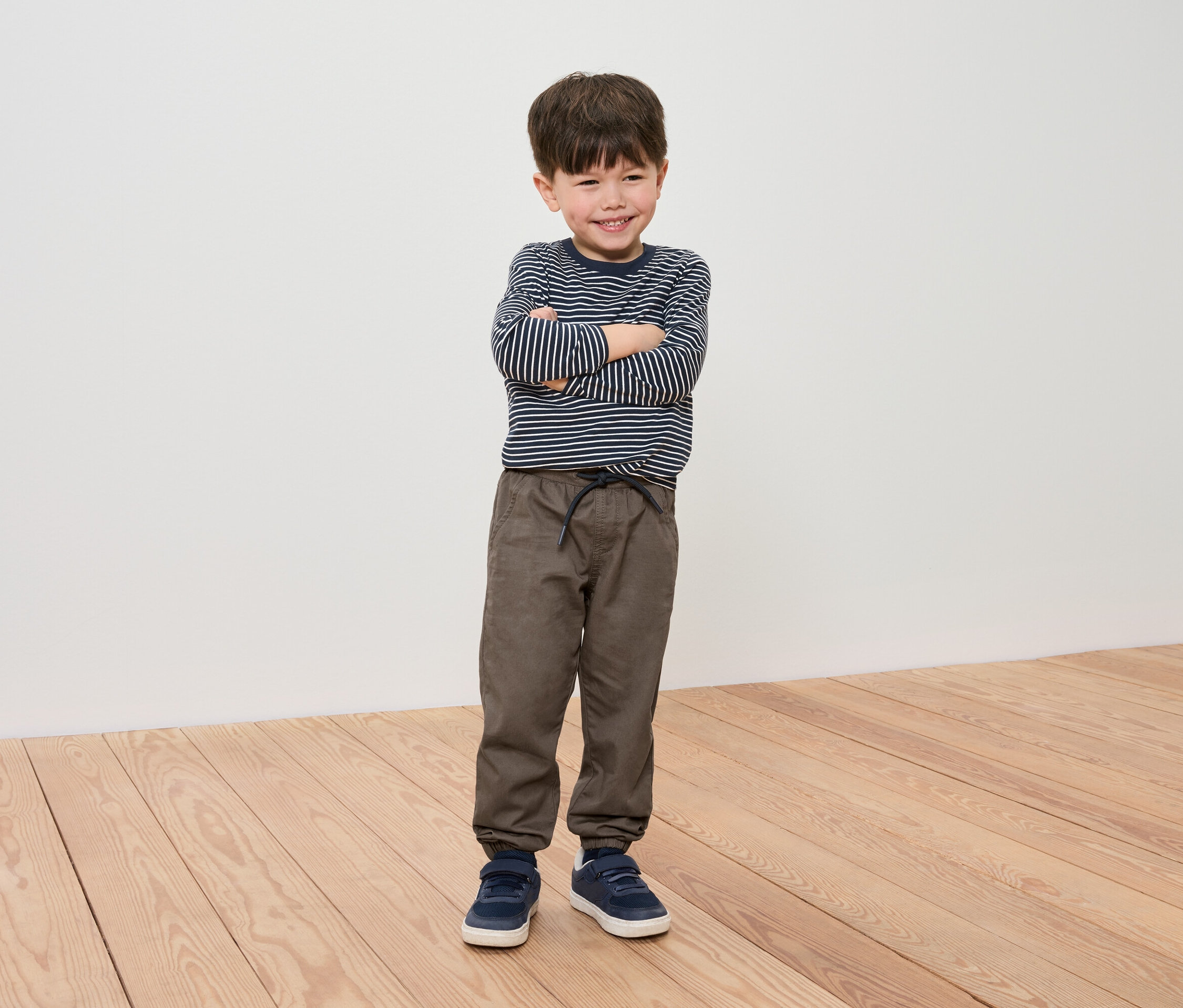 Ein Junge mit verschränkten Armen posiert in einem gestreiften Shirt, braunen Hosen und blauen Turnschuhen.