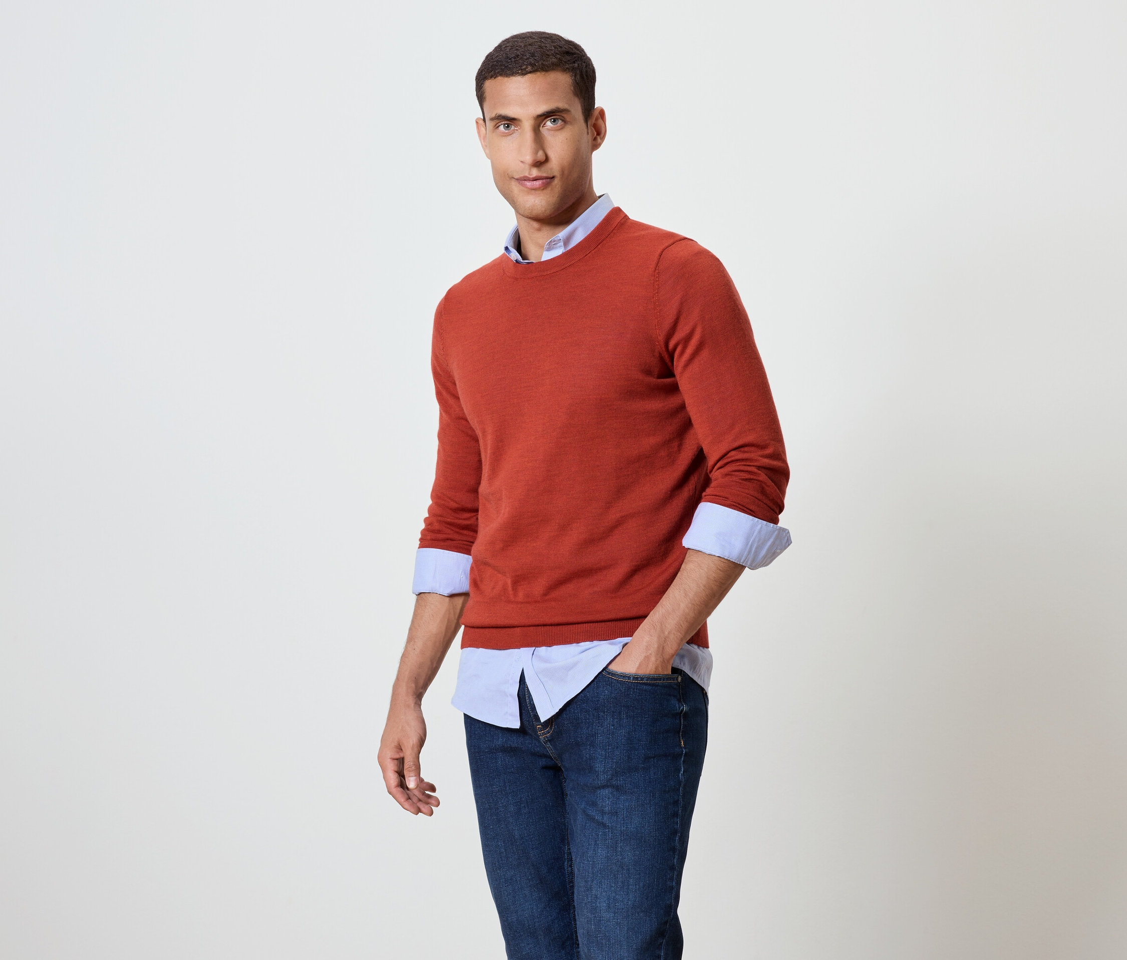 Mann posiert in orangefarbenem Pullover über blauem Hemd und Jeans.
