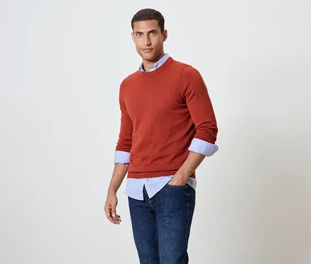 Mann posiert in orangefarbenem Pullover über blauem Hemd und Jeans.