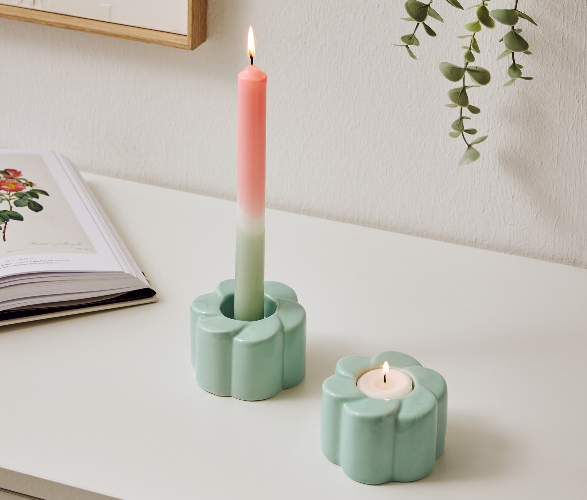 Zwei hellblaue Kerzenhalter im Blumen Design stehen auf einem Tisch, einer mit einer brennenden, zweifarbigen Kerze.