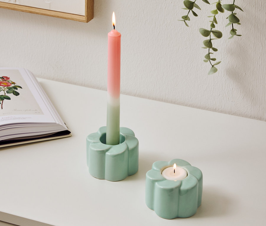 Zwei hellblaue Kerzenhalter im Blumen Design stehen auf einem Tisch, einer mit einer brennenden, zweifarbigen Kerze.