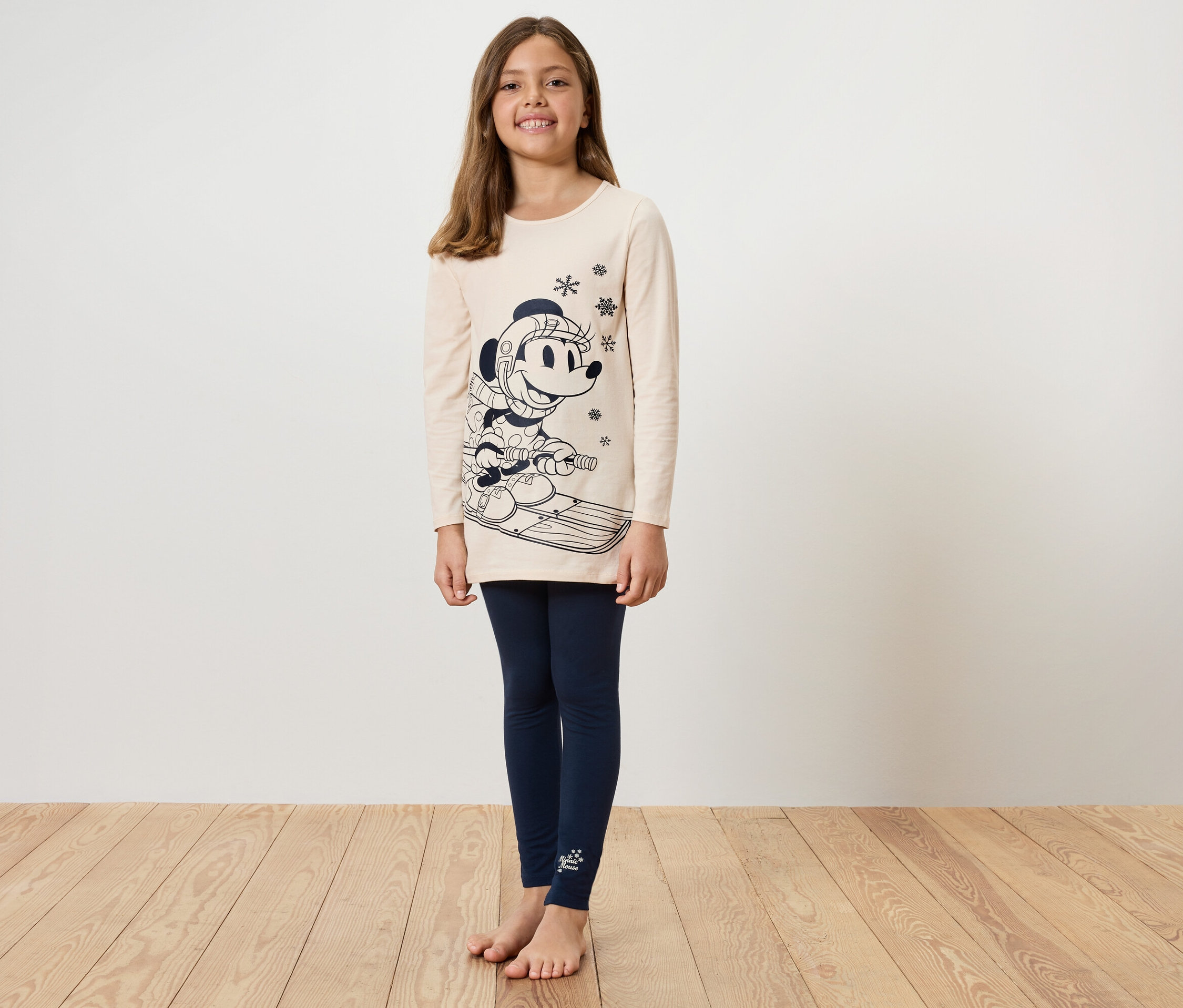 Mädchen in beigem Shirt mit Mickey Mouse Druck auf Skiern und blauen Leggings steht auf einem Holzboden.