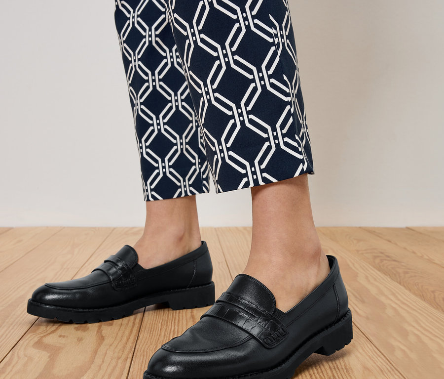 Beine einer Person in schwarzen Loafern und blauen gemusterten Hosen, die auf einem Holzboden stehen.