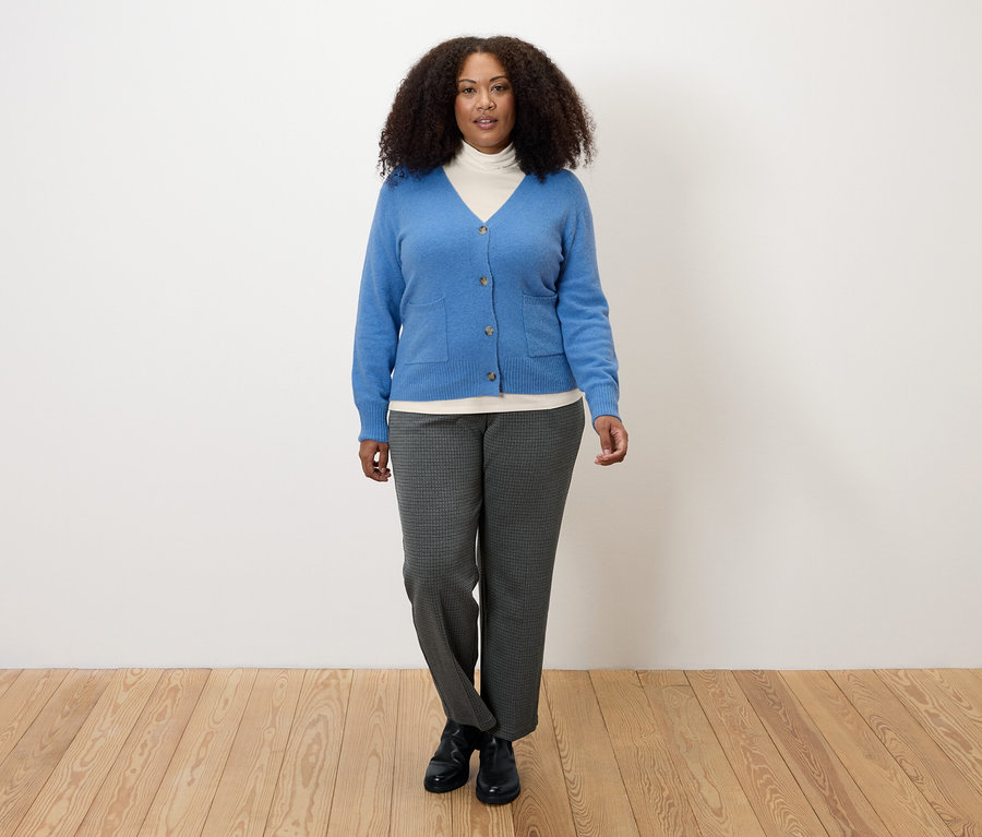 Frau trägt ein cremeweißes Rollkragenshirt, eine blaue Strickjacke und graue Hosen.