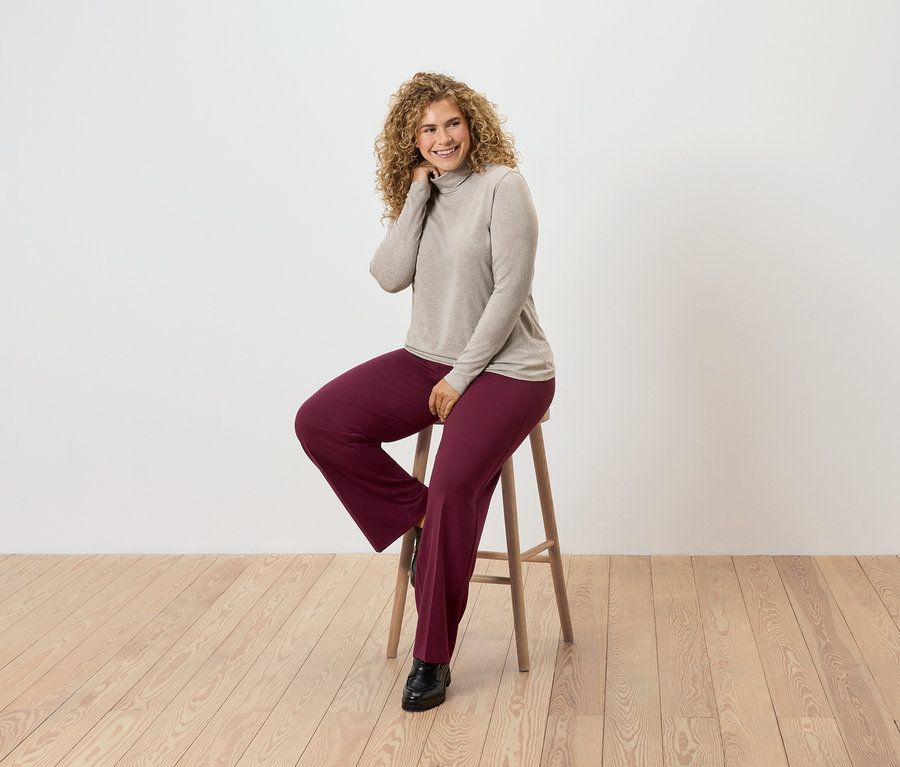Frau mit lockigem Haar sitzt auf einem Holzhocker, trägt einen beigen Rollkragenpullover und bordeauxrote Hosen.