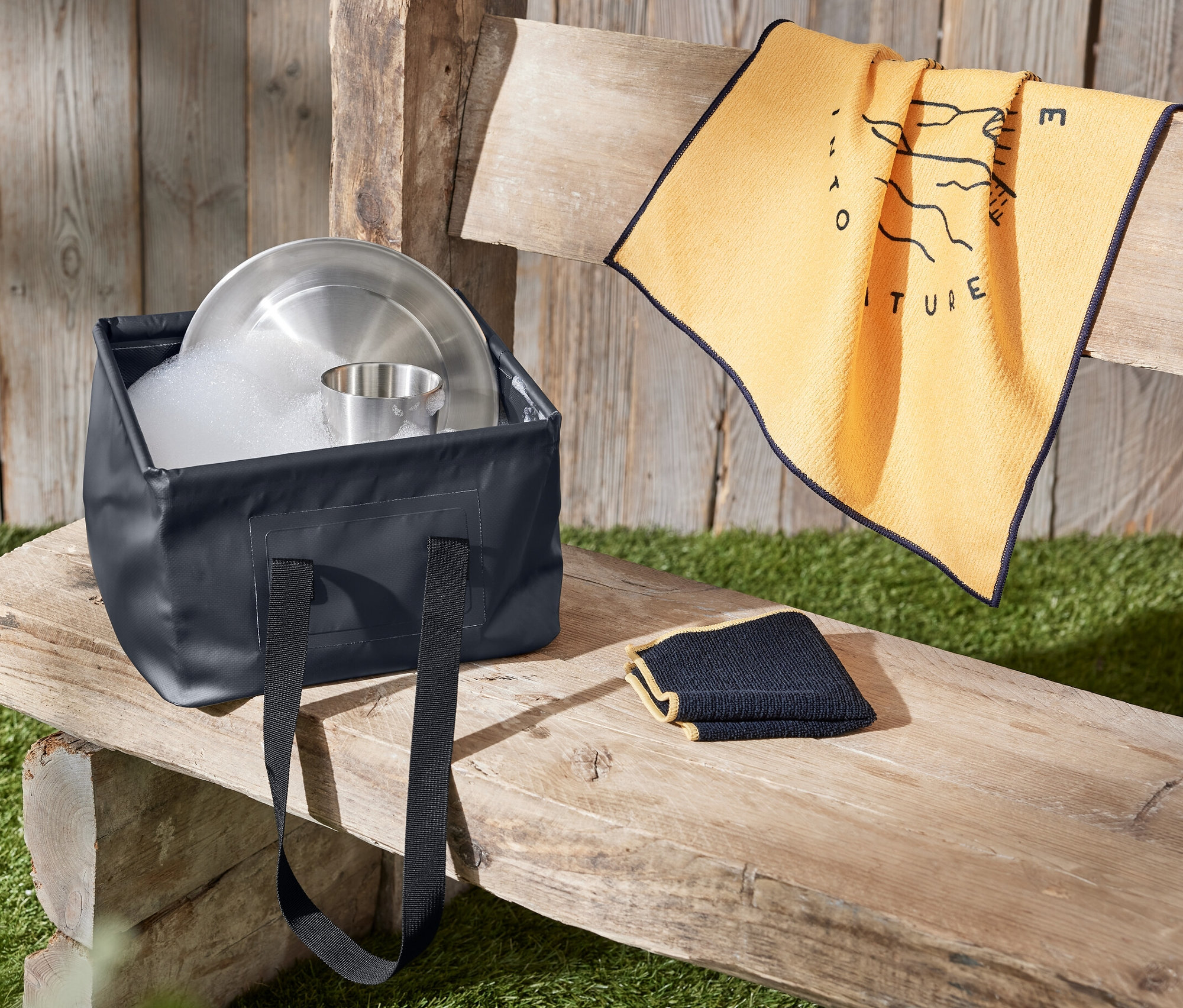 Eine faltbare Spülschüssel voller Wasser und Geschirr steht auf einer Holzbank neben einem Handtuch.