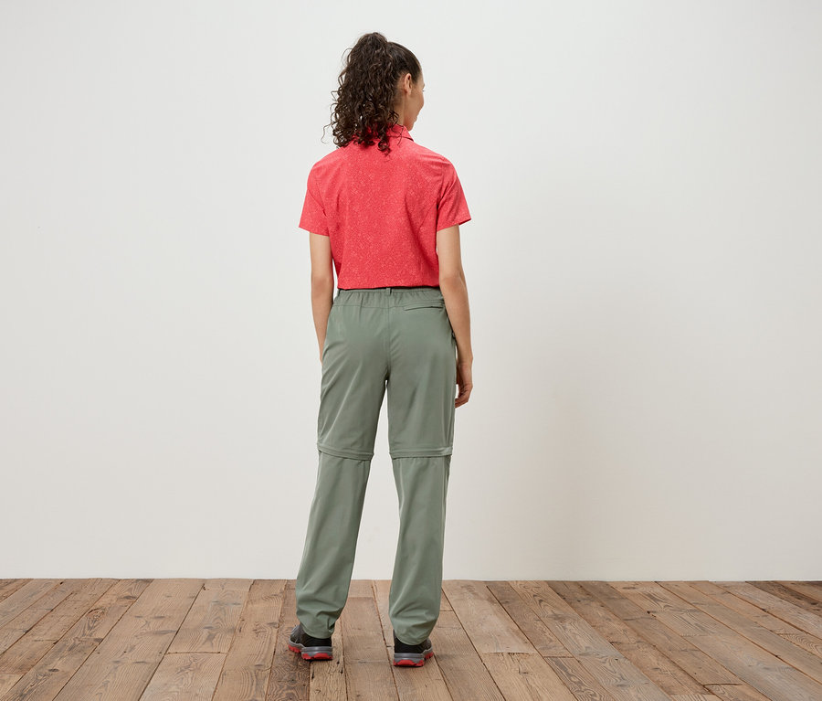 Rückansicht einer Frau mit lockigem Haar, die ein rotes Hemd und eine olivgrüne Zip-Off-Hose vor einem weißen Hintergrund trägt.