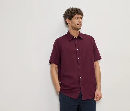 Mann mit lockigem Haar, der ein weinrotes, kurzärmeliges Hemd und eine marineblaue Hose vor einer weißen Wand trägt.