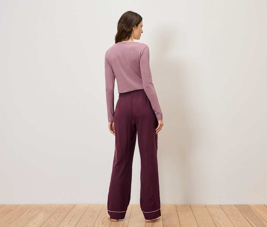Eine Frau mit langen, braunen Haaren trägt ein lila Oberteil und eine weinrote Hose mit weißem Saum.
