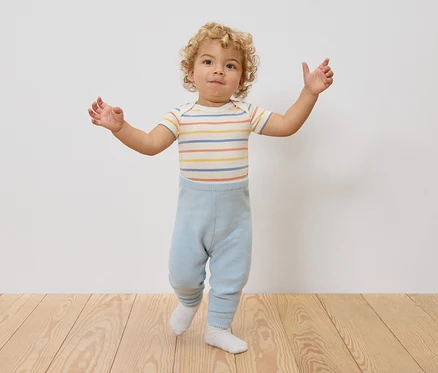 Ein Kleinkind mit lockigem Haar steht mit ausgestreckten Armen da, trägt einen gestreiften Body und hellblaue Leggings.