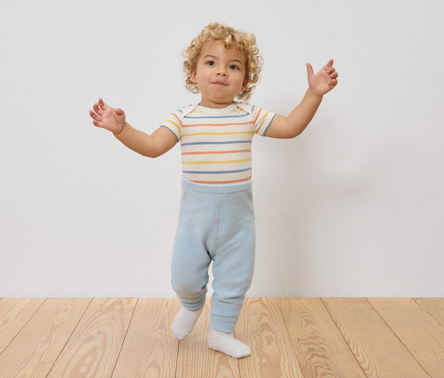 Ein Kleinkind mit lockigem Haar steht mit ausgestreckten Armen da, trägt einen gestreiften Body und hellblaue Leggings.
