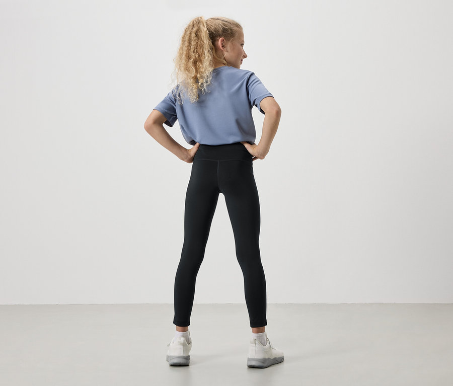 Rückansicht eines jungen Mädchens mit blonden, lockigen Haaren, das ein blaues T-Shirt, schwarze Leggings und weiße Turnschuhe trägt, die Hände in die Hüften gestemmt.