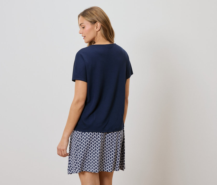 Rückansicht einer Frau mit blonden Haaren, die ein dunkelblaues T-Shirt und einen Rock mit blau-weißem Muster trägt.