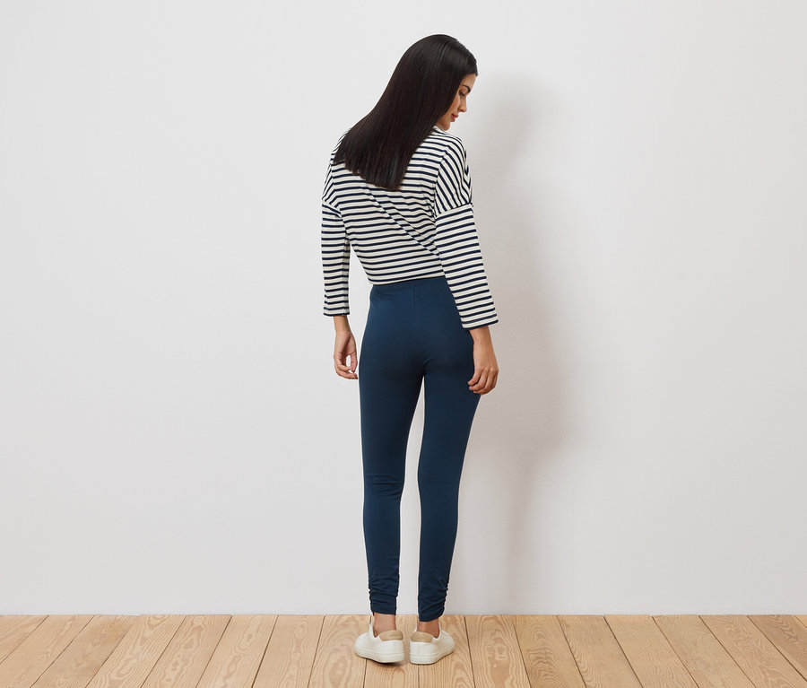 Eine Frau mit langen, dunklen Haaren steht mit dem Rücken zum Betrachter, trägt ein gestreiftes Oberteil, blaue Leggings und weiße Schuhe.