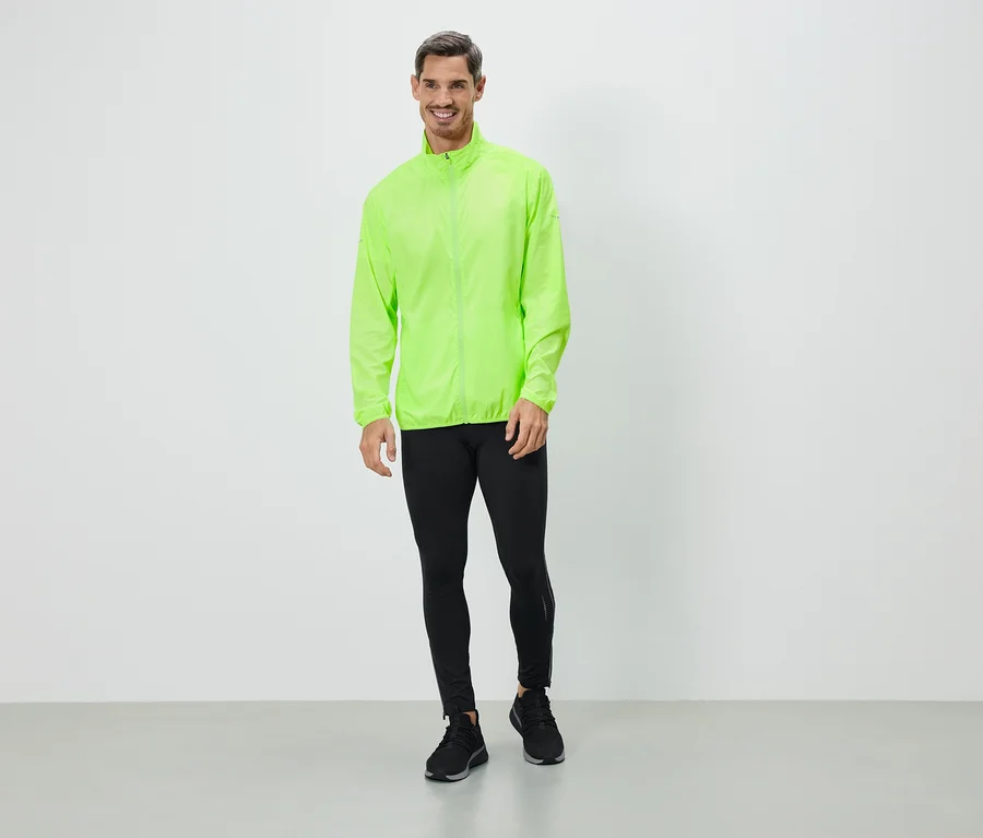 Mann in neongrüner Jacke, schwarzen Leggings und Turnschuhen steht vor einer weißen Wand.