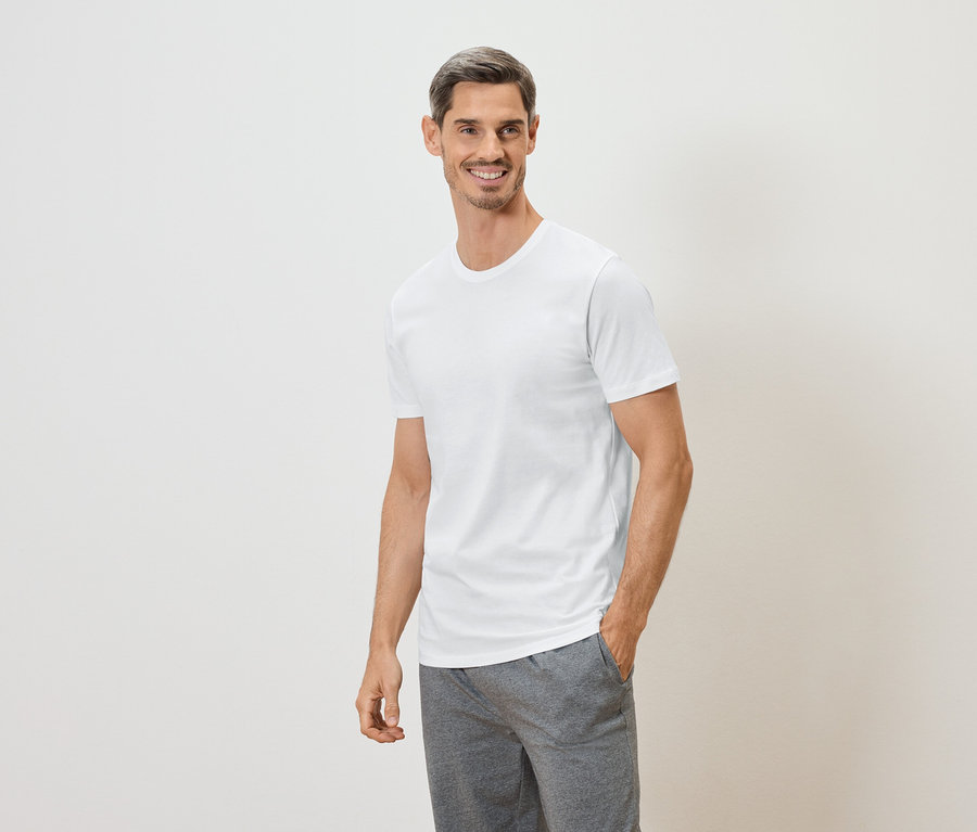 Lächelnder Mann mit Hand in der Hosentasche, trägt ein weißes T-Shirt und graue Shorts vor einem weißen Hintergrund.