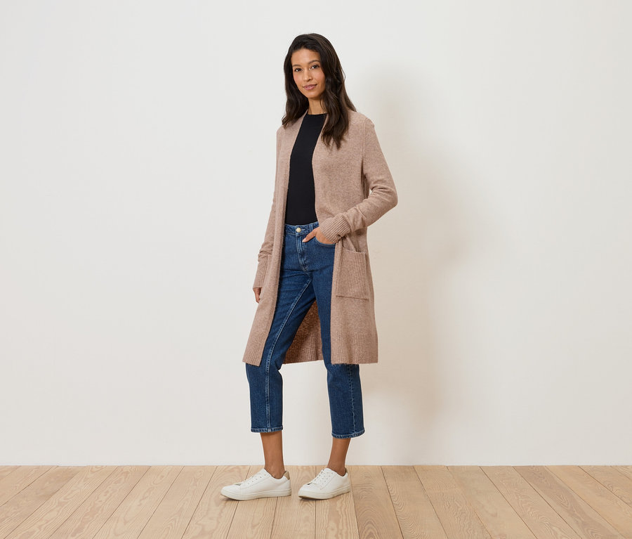 Frau steht in langem, beigefarbenem Cardigan, Jeans und weißen Turnschuhen vor einer weißen Wand.