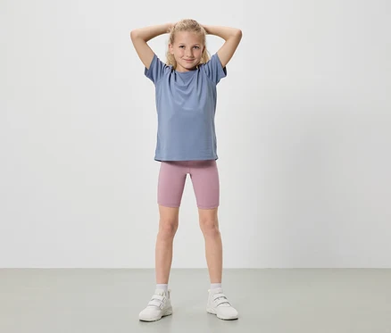 Mädchen mit blonden Haaren steht mit erhobenen Händen, trägt ein blaues T-Shirt, rosafarbene Radlerhosen und weiße Turnschuhe.