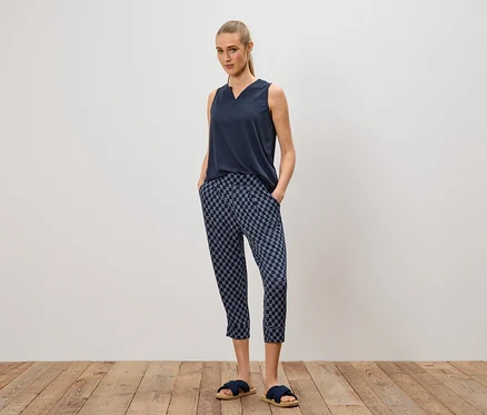 Frau steht mit Händen in den Hosentaschen, trägt ein marineblaues Top, gemusterte Hose und blaue Sandalen.