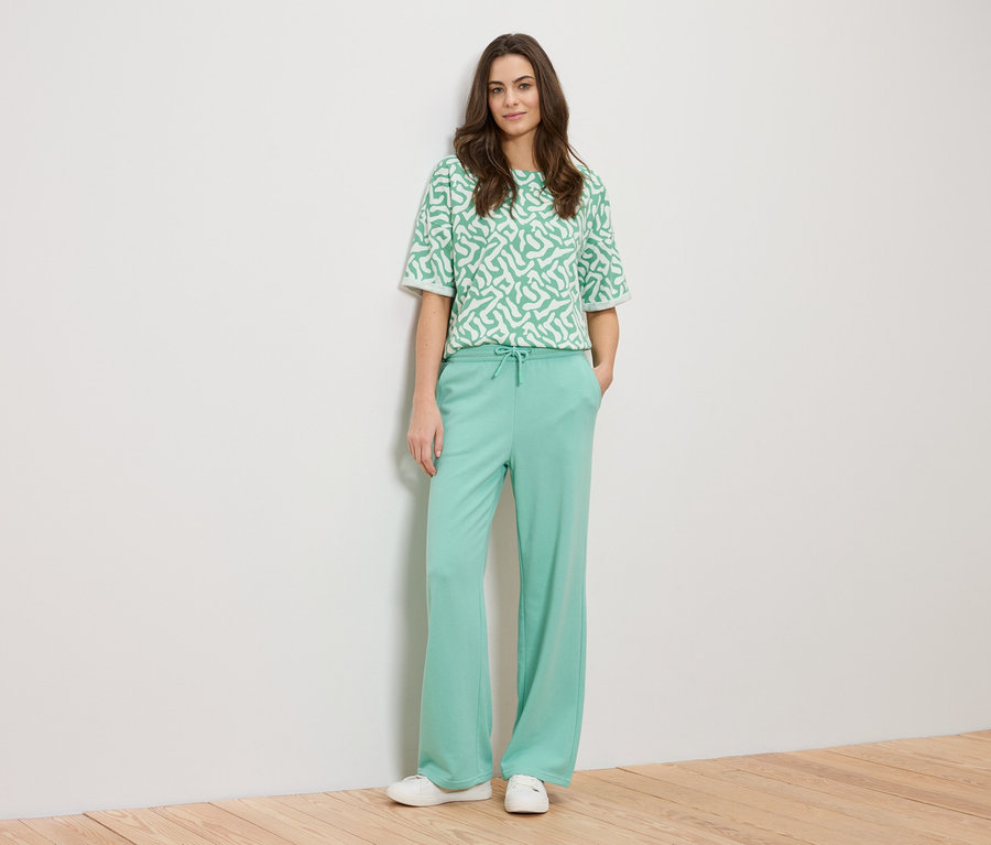 Eine Frau lehnt an einer Wand und trägt ein grünes, gemustertes Oberteil und eine grüne Hose.