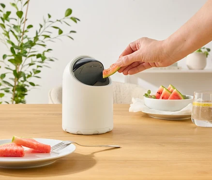 Eine Hand wirft ein Stück Wassermelone in einen weißen, runden Mülleimer auf einem Holztisch. Teller mit Melone und ein Glas Wasser.