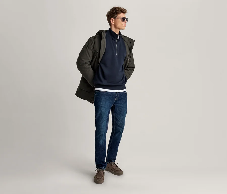 Mann trägt eine dunkelgrüne Parka über einem marineblauen Half-Zip-Sweatshirt, blaue Jeans, braune Schuhe und eine Sonnenbrille, blickt nach rechts.