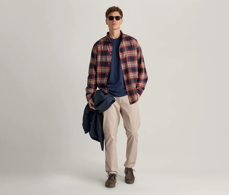 Mann trägt ein rot, marineblau und beige kariertes Hemd über einem marineblauen T-Shirt, beigefarbene Hosen und dunkelbraune Schuhe, hält eine marineblaue Jacke und trägt eine Sonnenbrille.