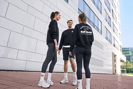 Drei Personen, zwei Frauen und ein Mann, stehen lächelnd und unterhalten sich. Alle tragen schwarze Sweatshirts und weiße Unisex Coffee Crew Tennissocken. Der Mann trägt schwarze Sportshorts. Eine Frau trägt grau-schwarze Sportleggings »ActivePower«, die andere schwarze Sportleggings »ActivePower«.
