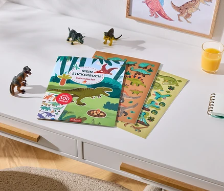 Ein weißer Schreibtisch mit einem Dinosaurier-Stickerbuch, das über 300 Sticker enthält, und zwei Stickerbögen. Daneben stehen drei kleine Dinosaurierfiguren, ein Glas Orangensaft, ein Notizbuch und zwei Buntstifte. Im Hintergrund hängt ein gerahmtes Dinosaurierbild an der Wand.