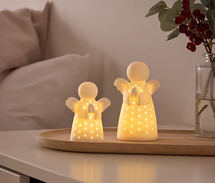 Zwei leuchtende, weiße Engelsfiguren mit ausgestanzten Löchern stehen auf einem Holztablett auf einem Nachttisch. Im Hintergrund sind ein Bett und eine Vase mit roten Beeren zu sehen.