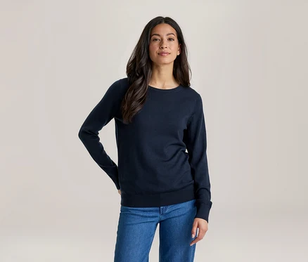 Eine Frau mit langen dunklen Haaren, die ein dunkelblaues Langarmshirt und blaue Jeans trägt, steht mit einer Hand in der Hüfte und blickt nach vorne.