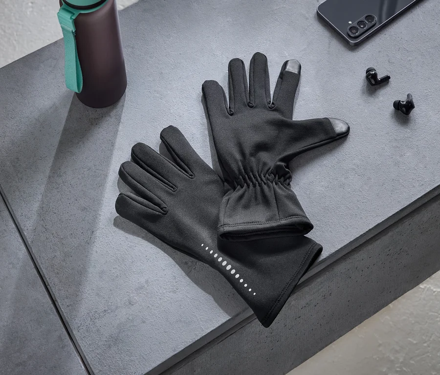 Ein Paar schwarze Handschuhe mit reflektierenden Punkten und Touchscreen-kompatiblen Fingerspitzen, eine dunkle Trinkflasche mit einem türkisen Riemen, ein schwarzes Smartphone und schwarze kabellose Kopfhörer liegen auf einer grauen Oberfläche.