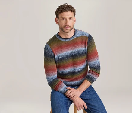 Mann sitzt auf einem Hocker und trägt einen bunten, gestreiften Rundhalspullover in Rot-, Blau-, Grün- und Brauntönen sowie blaue Jeans.