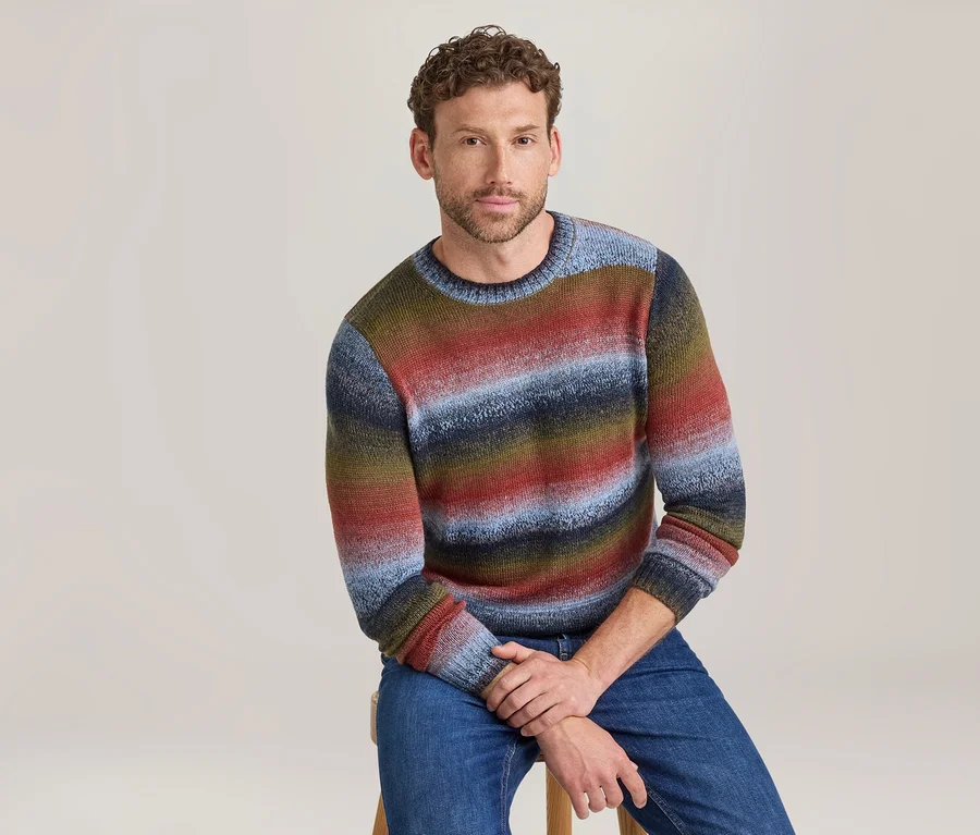 Mann sitzt auf einem Hocker und trägt einen bunten, gestreiften Rundhalspullover in Rot-, Blau-, Grün- und Brauntönen sowie blaue Jeans.