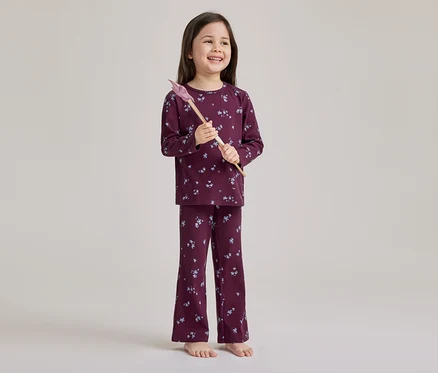 Ein lächelndes junges Mädchen steht barfuß und trägt einen gerippten, bordeauxfarbenen Pyjama mit hellem Blumenmuster. Sie hält einen rosa-braunen Spielzeugzauberstab.
