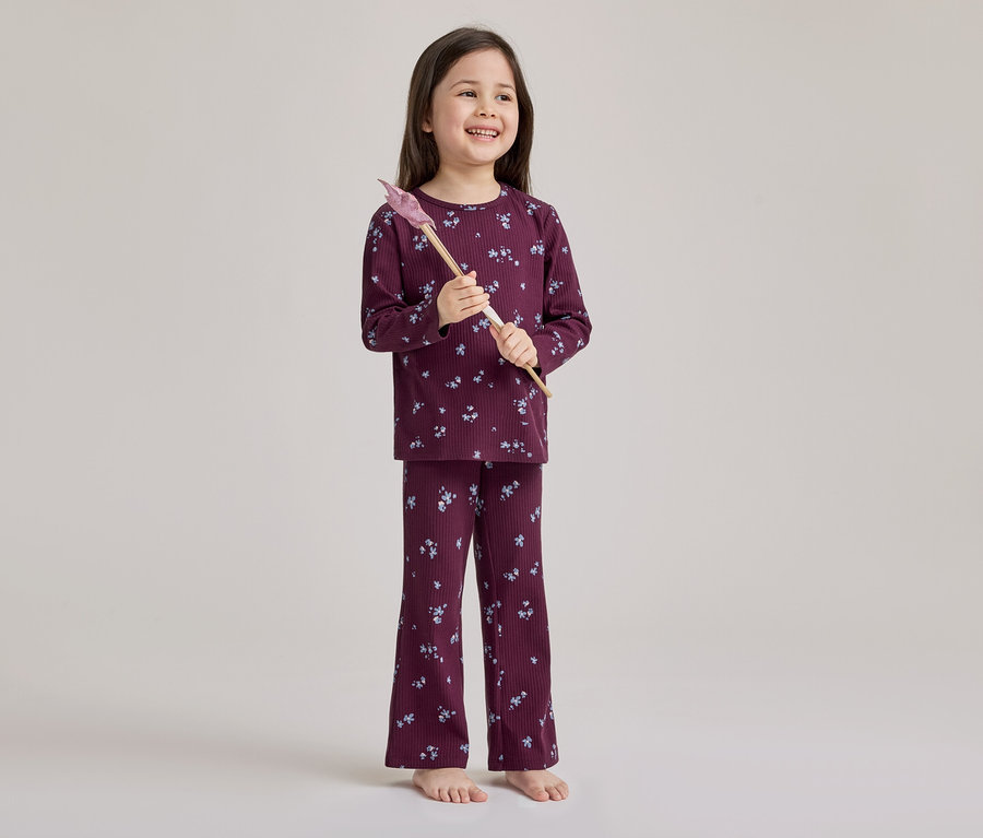 Ein lächelndes junges Mädchen steht barfuß und trägt einen gerippten, bordeauxfarbenen Pyjama mit hellem Blumenmuster. Sie hält einen rosa-braunen Spielzeugzauberstab.