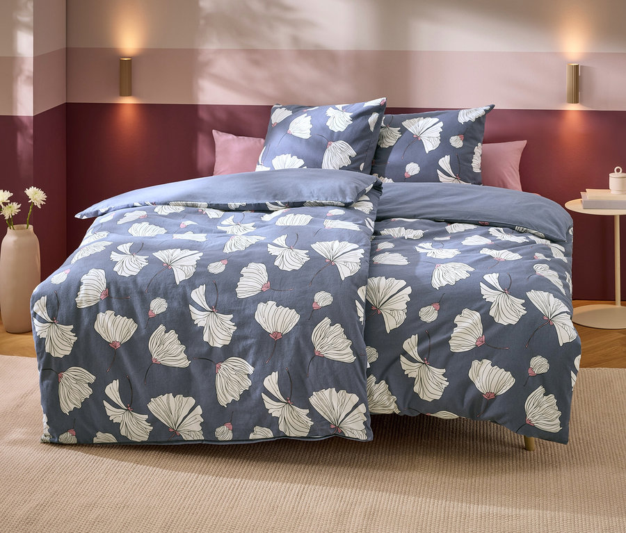 Ein Bett mit blauer Bettwäsche mit weißen Blumenmustern und rosa Akzenten. Dahinter zwei rosa Kissen. Die Wand ist zweifarbig lila, mit zwei goldenen Wandleuchten. Links steht eine Vase mit weißen Blumen, rechts ein Beistelltisch mit Büchern.