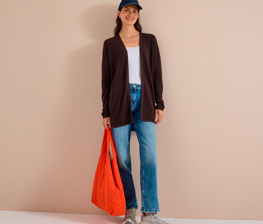 Frau steht lächelnd, trägt eine dunkelbraune CECIL Strickjacke, weißes Top, Jeans, blaue Kappe und hält eine orangefarbene Stepptasche.