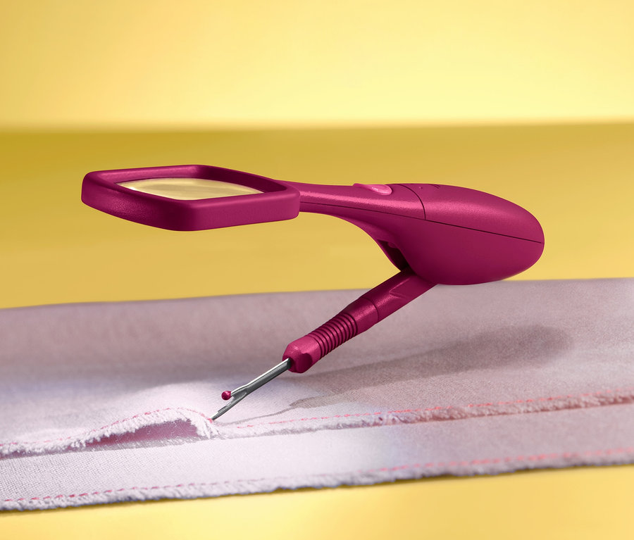 Nahaufnahme eines pinkfarbenen Nahttrenners mit Lupe und LED-Licht in Verwendung auf einem Stoff mit rotem Faden vor gelbem Hintergrund.