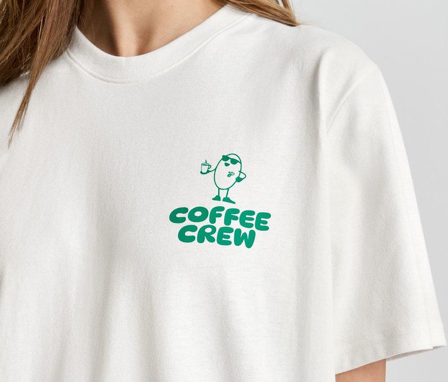 Nahaufnahme einer Person, die ein helles T-Shirt mit dem grünen Aufdruck "Coffee Crew" und einer Cartoon-Kaffeebohne trägt.